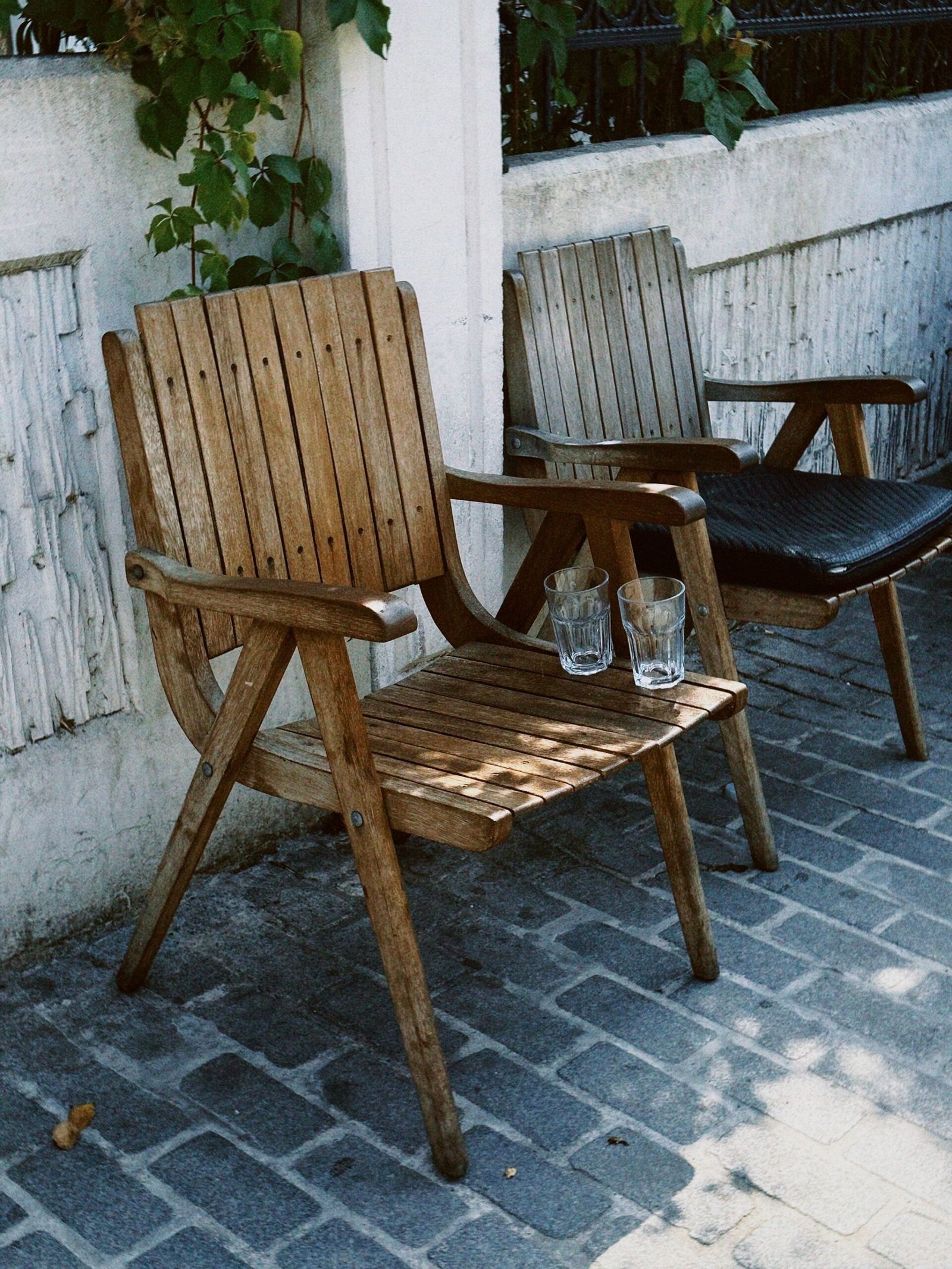 Vintage wooden chairs on a patio with two empty glasses on a sunny day.
